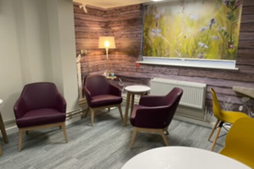 A room with yellow dining chairs, wooden tables, grey wood effect floor and purple coloured armchairs. Wall art on the rear wall depicts soft focus flowers and grasses which co-ordinate with the furniture. There is a lamp switched on in the corner.