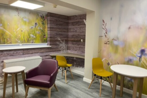 A room with yellow chairs, wooden tables, grey wood effect floor and purple coloured armchairs. Wall art on the rear wall depicts soft focus flowers and grasses which co-ordinate with the furniture.