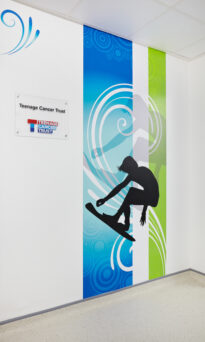 A wall with a Teenage Cancer Trust logo  and a silhouette of a person on a surfboard with blue and green swirls in the background.