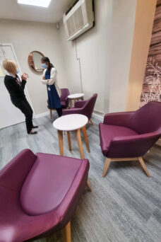 Two ladies are chatting in a room with grey wood effect floor, several purple easy chairs and a small wooden table. The edge of wall art featuring light purple wood effect with silhouetted grasses is visible to the right of the picture.