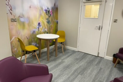 A room with yellow chairs, a wooden table, grey wood effect floor and a mural and purple coloured armchairs. Wall art on the rear wall depicts soft focus flowers and grasses which co-ordinate with the furniture.