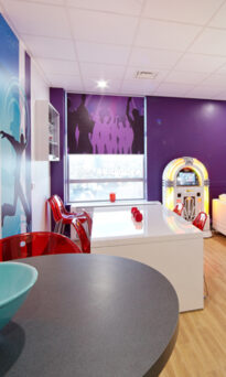 A room with purple and turquoise wall art depicting concert goers.  There is a juke box and white furniture in the room with co-ordinating chairs in a pinky red and a round table.